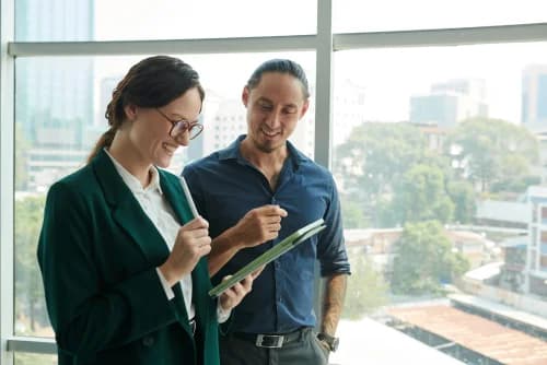 Dos profesionales revisan información en una tablet junto a un ventanal con vista a la ciudad