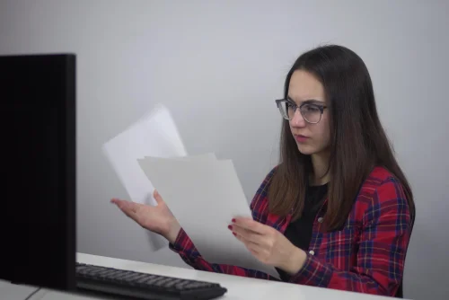 Mujer con gafas revisa unas hojas impresas sentada frente a un computador